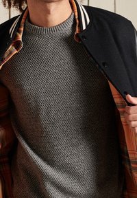 Person in einem strukturierten grauen Strickpullover und einer schwarzen Jacke mit gestreiftem Kragen, die eine orange-schwarze Karolierung zeigt.