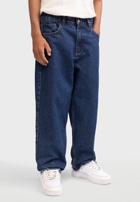 Jean en denim bleu avec une coupe décontractée, avec cinq poches, une fermeture à bouton et des coutures contrastées, associés à des baskets blanches.