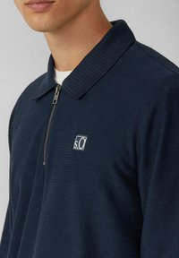 Navy Pullover mit geripptem Struktur, Reißverschluss und flachem Kragen. Mit einem weißen gestickten Logo auf der Brust.