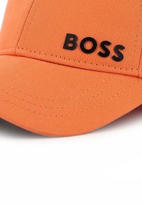 Chapeau orange avec un bord incurvé, fabriqué en tissu texturé, arborant un logo noir « BOSS » à l'avant. Couture décorative sur le bord.