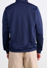 Mies, joka käyttää tummansinistä takkia ja vaaleita housuja, näkyy selkäpuolelta yksinkertaista vaaleaa taustaa vasten.