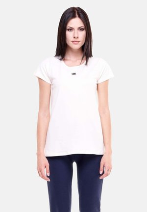 Giovane donna con capelli lisci e scuri, che indossa una maglietta bianca semplice e pantaloni scuri, in piedi davanti a uno sfondo chiaro e uniforme.