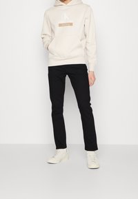 Sudadera con capucha de color crema con el logo de Calvin Klein Jeans, combinada con jeans ajustados negros y zapatillas blancas. Tejido suave, diseño relajado.