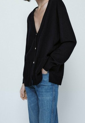 Osoba nosząca czarny rozpinany kardigan z długimi rękawami i dwoma przednimi kieszeniami, w połączeniu z niebieskimi dżinsami, na tle jednolitego jasnego tła.