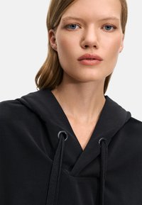 Hanorac negru cu un design tip V, având un sistem de închidere cu șnur și un material moale, texturat. Stil minimalist și culoare neutră.
