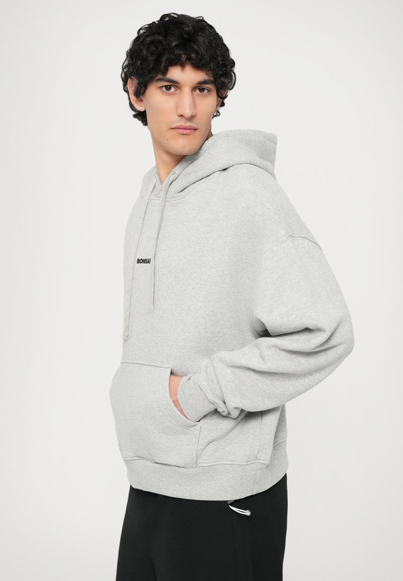 Gray hooded sweatshirt with a kangaroo pocket and drawstring, featuring a small black logo on the chest. Soft, textured fabric.