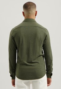 Man met kort haar, gekleed in een olijfgroene lange mouwen shirt en witte broek, staande met zijn rug naar de camera tegen een effen achtergrond.