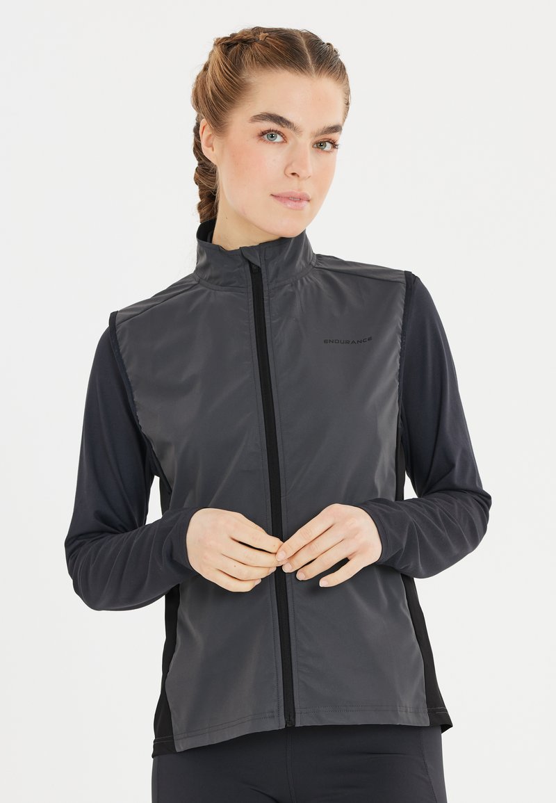 Schwarze ärmellose Jacke mit hohem Kragen, ausgestattet mit einem durchgehenden Reißverschluss und kontrastierenden schwarzen Seitenpaneelen. Glatte, glänzende Stofftextur.