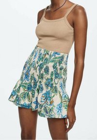 Beige gerippter Tanktop kombiniert mit einem floral gemusterten, gesmokten Minirock, der blaue und grüne Blumen auf einem hellen Hintergrund zeigt, mit rüschensaum.