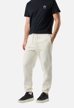 Homme portant un t-shirt noir, un pantalon de jogging écru avec cordon de serrage et des baskets noires avec semelles blanches, debout avec une main dans la poche.