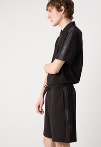 Polo shirt nera e pantaloni corti coordinati realizzati in un tessuto liscio. La maglietta presenta maniche corte con dettagli grigi; i pantaloni hanno una vita con cordino.