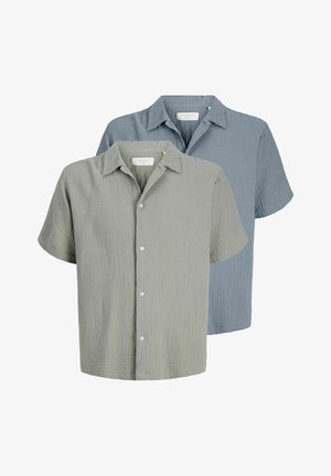 Ung mann iført lysegrønn kortermet knappeskjorte, mørke vide denimshorts, hvite sokker og beige sko, stående mot ensfarget bakgrunn.