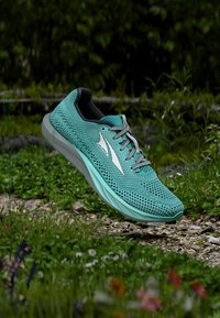 Lätt löparsko med övre del i teal-mesh, med grå accent och vadderad sula, svävande ovanför gräs och stenar.