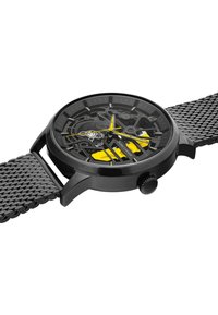 Schwarze Mesh-Armbanduhr mit einem transparenten schwarzen Zifferblatt, das gelbe Akzente und komplexe mechanische Details aufweist. Rundes Gehäuse mit texturiertem Krone.