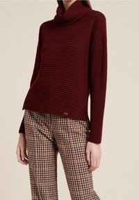 Burgundy ribbad tröja med hög hals, kombinerad med bruna och orange houndstooth-mönstrade byxor. Accenterad med en liten logodetalj.