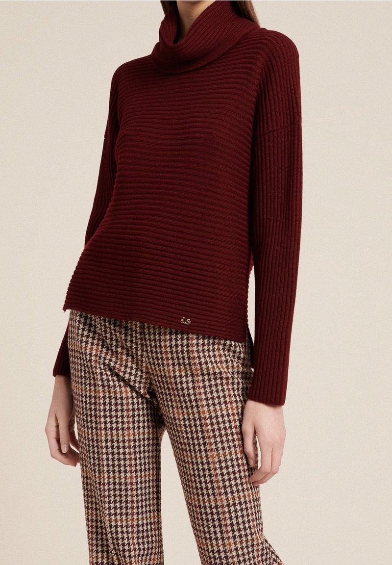 Burgundy ribbad tröja med hög hals, kombinerad med bruna och orange houndstooth-mönstrade byxor. Accenterad med en liten logodetalj.