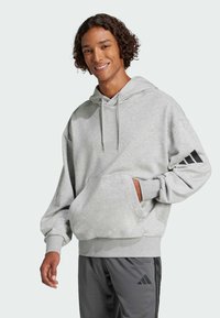 Felpa grigia con cappuccio, tasca a marsupio, vestibilità rilassata e dettagli neri del logo Adidas sulle maniche. Tessuto morbido e con texture.