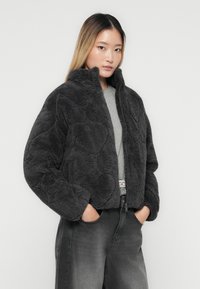 Kvinde med langt glat hår iført en mørkegrå fluffy zip-up jakke, lys grå skjorte og slidte sorte jeans med hænderne i lommerne.