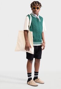 Grüner Strickwesten über einem weißen T-Shirt, schwarze Shorts, braune Slipper, gestreifte Socken, hält eine cremefarbene Tragetasche, mit Sonnenbrille und einem Schal.