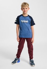 Blå och marinblå kortärmad t-shirt med vit logotyp, kombinerad med vinröda mjukisbyxor och grå sneakers med blå detaljer.