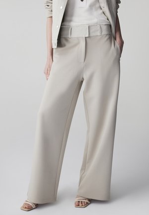 Persona che indossa pantaloni beige a gamba larga, top bianco a coste e una giacca beige chiaro con una mano in tasca, in piedi su uno sfondo neutro.