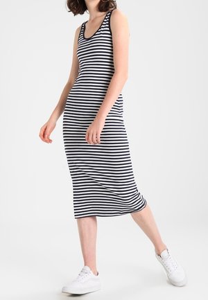 Woman wearing a black and white striped sleeveless midi dress and white sneakers, standing with one leg slightly lifted.