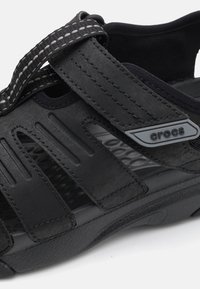 Czarne sandały z syntetycznej górnej części z otworami wentylacyjnymi, teksturowaną podeszwą i paskiem z odblaskowym przeszyciem. Obecne logo Crocs.