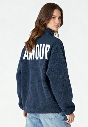 Pull polaire bleu marine à col montant, avec un grand texte blanc "AMOUR" au dos. Texture douce, coupe décontractée, épaules tombantes.