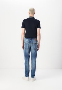 Polo shirt bleumarin cu margini albe, asortat cu blugi deschiși, deteriorați, și adidași albi. Persoana este prezentată din spate, pe un fundal minimalist.