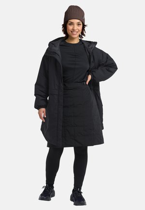 Black puffer coat with a zippered front, paired with a fitted black top, knee-length skirt, and black leggings. Ribbed brown beanie on head.