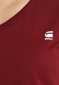 Närbild av en person som bär en vinröd skjorta med en vit "RAW"-logotyp tryckt nära halsringningen på vänster sida.