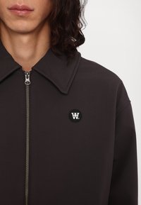 Jachetă maro cu fermoar și guler lat, având o patch cu logo rotund negru. Textură moale și detalii cu fermoar aurii.