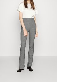 Mujer vistiendo pantalones acampanados en patrón de jaquard blanco y negro, camisa blanca de manga corta y botines negros de tacón, de pie contra un fondo liso.