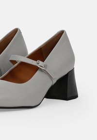 Zapatos Mary Jane de cuero gris con punta redondeada, correa ajustable con hebilla plateada y tacón cuadrado negro texturizado.