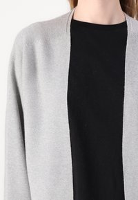 Světle šedý cardigan vyrobený z pletené látky, s jemnou texturou a otevřeným designem vpředu. Nesený přes černý top, který ukazuje kontrastní barvy.