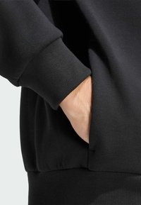 Čierna mikina so širším patentováním na rukávoch a spodnom leme, s rukou odloženou v bočnej vrecku, predvádzajúca hladkú textúru látky.