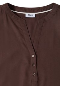Blouse marron foncé avec un col rond, une patte de boutonnage partielle à quatre boutons, et une étiquette de marque "CECIL" à l'intérieur du col.