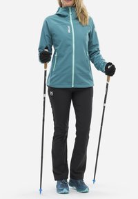 Türkise Softshelljacke mit hellen Akzenten, schwarze Hose und blaue Wanderschuhe. Verwendet Trekkingstöcke und trägt Handschuhe.
