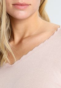 Haut rose clair avec un col en V festonné, en tissu côtelé, montrant une texture douce et des détails de surface lisse.