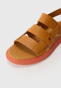 Brune lærsandaler med tre brede remmer, flad sål og struktureret orange gummisål. Syningsdetaljer og små metalaccent.