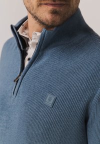 Blue knit pullover with a zipper collar and ribbed texture. Features a logo patch and a contrasting collared shirt underneath.