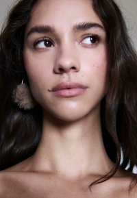 Primo piano di un volto con pelle liscia. Le caratteristiche includono occhi marrone scuro, labbra naturali e un orecchino peloso di colore marrone chiaro. Capelli scuri e mossi.