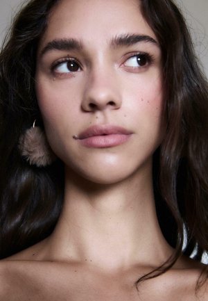 Zbliżenie twarzy z gładką skórą. Cechy to ciemnobrązowe oczy, naturalne usta i futrzane, jasnobrązowe kolczyki. Ciemne, faliste włosy.