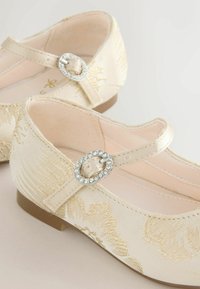 Cremefarbene Stoffschuhe mit floralem Muster, Riemendetail mit einer Schnalle besetzt mit Strasssteinen, flache Sohle und leicht strukturierte Oberfläche.