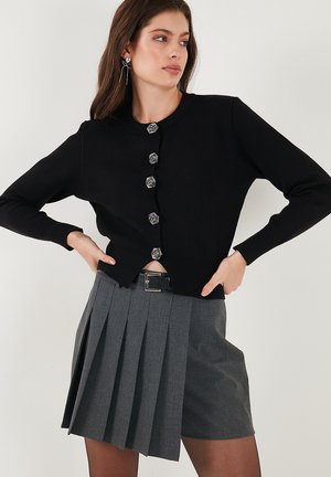 Black knitted cardigan with round neckline and silver floral buttons. Paired with gray pleated shorts and a black belt. Textured fabric details.