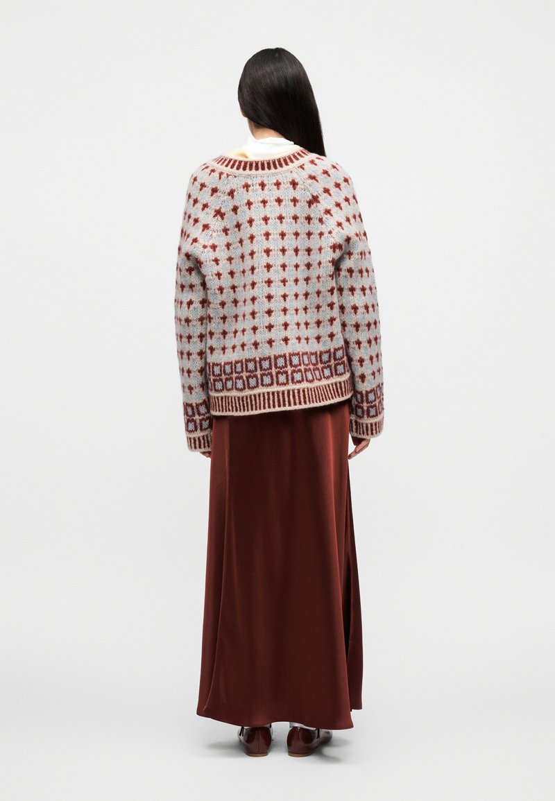 Frau, die mit dem Rücken zur Kamera steht, trägt einen gemusterten Wollpullover, einen langen rostfarbenen Rock und dunkelbraune Schuhe vor weißem Hintergrund.