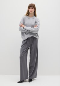Hellgrauer Strickpullover mit Rundhalsausschnitt, getragen über locker sitzenden grauen Hosen. Das Model steht mit verschränkten Armen und trägt schwarze spitze High Heels.