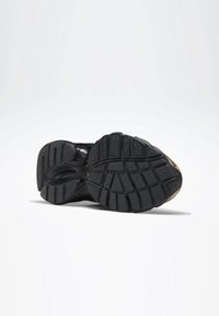 Paire de chaussures de sport à semelle noire positionnées semelle vers le haut, montrant des motifs de bande de roulement détaillés et une adhérence texturée sur un fond clair uni.