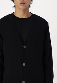Svart cardigan laget av strikket stoff, med dyp V-hals, fem knapper og lange ermer; glatt tekstur med subtile ribbede detaljer.