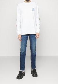Camicia bianca a maniche lunghe con testo grafico blu sul petto e design colorati sulle maniche, abbinata a jeans in denim blu e scarpe nere.